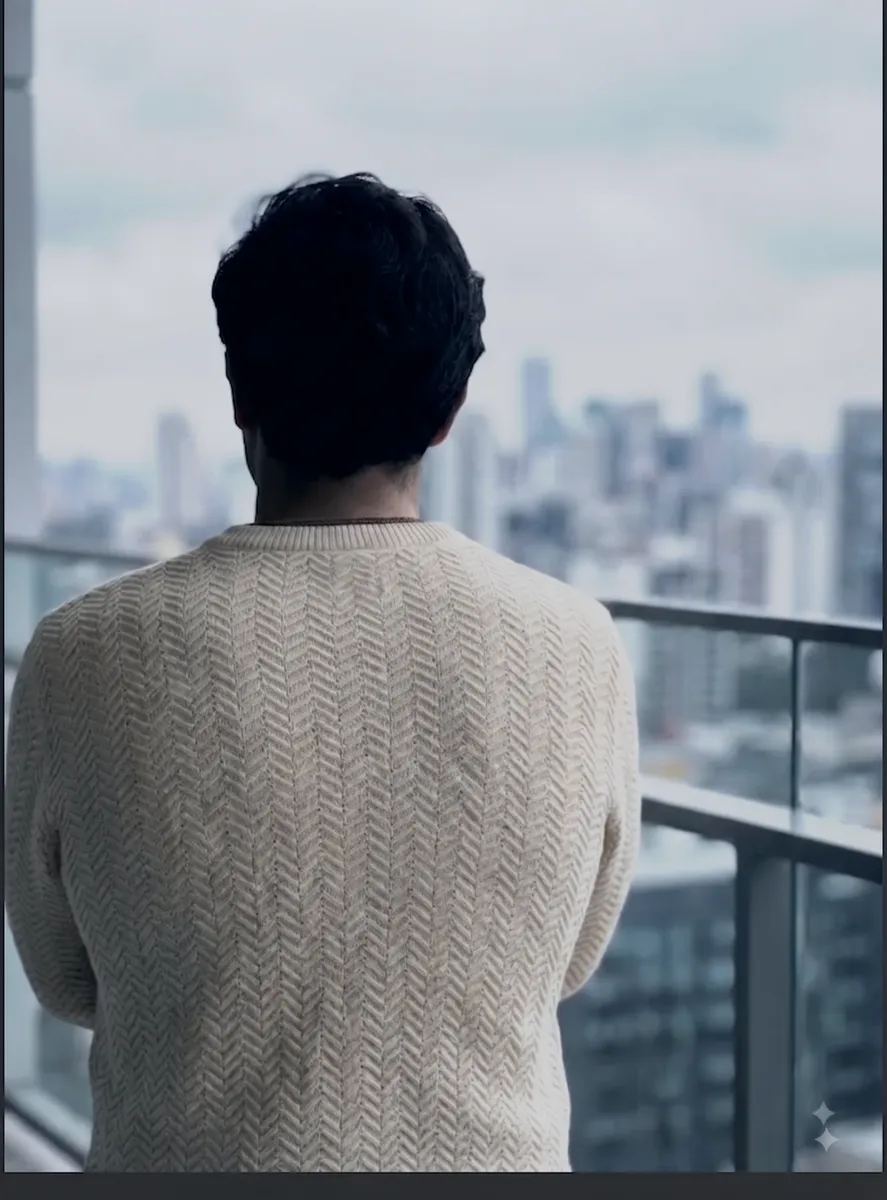 Modelo de costas com suéter de tricô cru em ponto origami, numa varanda com o skyline da cidade.