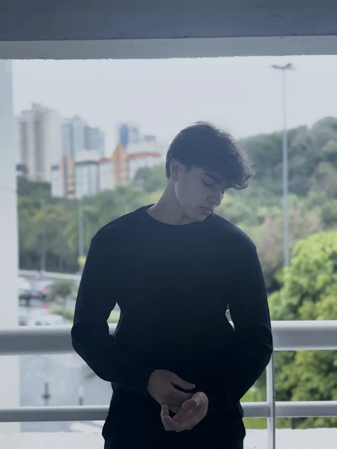 Modelo de suéter preto de tricô olhando para baixo numa varanda, prédios ao fundo.